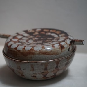 Puede incluir: Un tazón de cerámica blanco y marrón con tapa. El tazón y la tapa tienen un patrón único de puntos blancos sobre un fondo marrón.