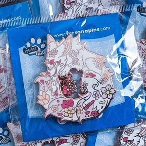 Op de afbeelding: Vijf witte emaillepins met een roze en wit bloemenontwerp. De pins tonen een witte vos met roze accenten en een roze bloem in zijn bek. De pins zijn verpakt in doorzichtige plastic zakjes met blauwe achterkaarten waarop "Fursona Pins" en "www.fursanapins.com" staat.