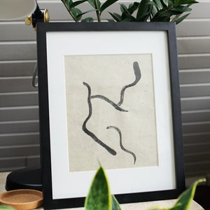 Puede incluir: Un marco negro con una impresión de arte abstracto y un passe-partout blanco. La obra presenta líneas negras fluidas sobre un fondo crema. Los tallos verdes de las plantas emergen de la parte superior del marco.
