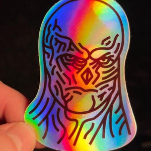 May include: A holographic sticker with a black and white line drawing of a face with a serious expression. The face is outlined in black and has a rainbow holographic background.