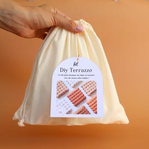 May include: A cream-coloured drawstring bag with a white tag that reads "DIY Terrazzo." The tag features a graphic of small, square terrazzo tiles in shades of white, pink, and coral. The bag is held against an orange background.