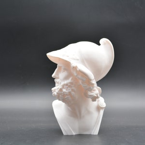 Pode incluir: Busto de gesso branco de um homem usando um capacete. O busto é uma vista de perfil do rosto e dos ombros do homem. O homem tem barba e cabelo comprido.
