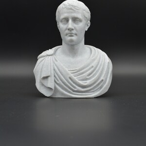 Puede incluir: Un busto de yeso blanco de un hombre con el pelo corto y una toga. El busto est&aacute; sobre un fondo negro.