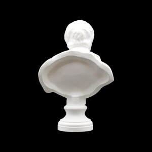 May include: A white bust sculpture, viewed from the back, set against a black background. The sculpture features a detailed depiction of a person's head and shoulders, with a classic, neoclassical style. The bust is mounted on a pedestal.