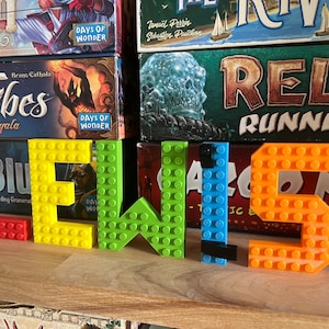 Può includere: Il nome "LEWIS" scritto con blocchi Lego colorati. I blocchi sono rosso, giallo, verde, blu e arancione. I blocchi sono su uno scaffale di legno davanti a una libreria con giochi da tavolo.