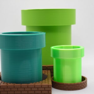 Puede incluir: Tres tuberías de plástico verde y verde azulado con bases de ladrillo marrón. Las tuberías son de diferentes tamaños y están apiladas una encima de la otra.