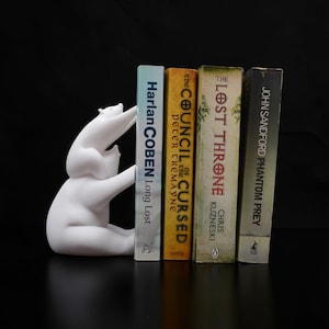 May include: A white polar bear bookend sculpture holding four books. The books have titles including "Long Lost", "The Council of the Cursed", "The Lost Throne", and "Phantom Prey". The bookend is on a black surface.