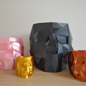 Peut inclure: Quatre pots de fleurs en forme de crâne géométrique de différentes tailles. Le plus grand pot est noir, le suivant est rose, le plus petit est doré et le plus petit est en cuivre.