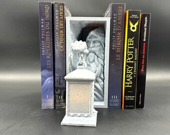 May include: A gray stone bookend featuring a wizard holding a lantern. The bookend is sitting on a shelf with several books, including "Les Royaumes du Nord", "La Tour des Anges", "Le Miroir d'Ambre", "Harry Potter et l'Enfant Maudit", and "Mary Ann en automne".