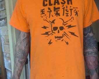 Può includere: Maglietta arancione con una grafica nera di teschio e ossa incrociate. Il testo "The Clash" e caratteri giapponesi sono stampati sulla maglietta.