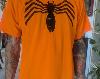 Può includere: Una maglietta arancione brillante con un design a ragno nero del simbionte Venom stampato sul davanti.