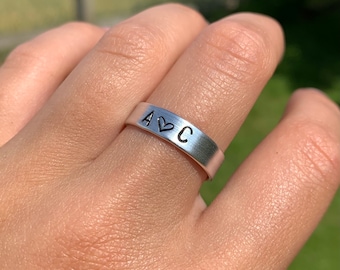 May include: A silver ring with the letters A and C engraved on it, separated by a heart symbol.