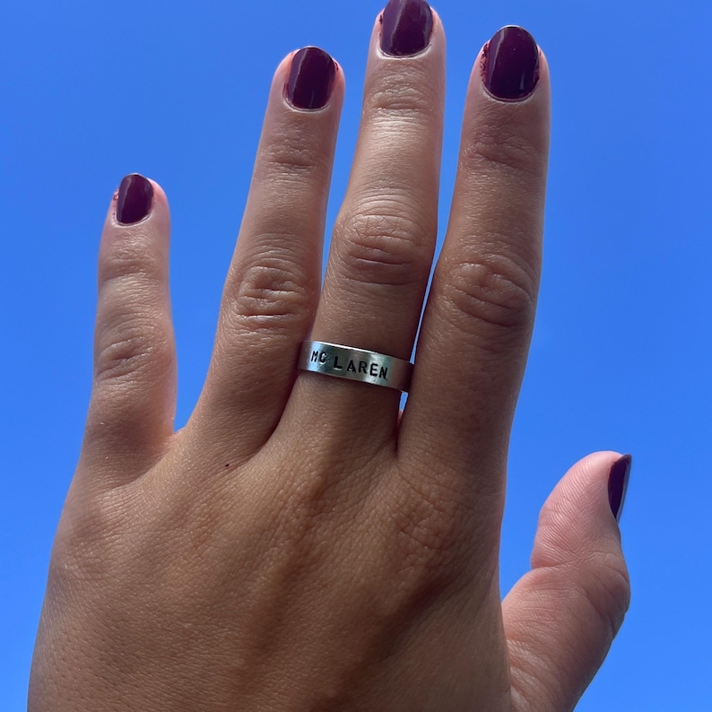 Puede incluir: Un anillo plateado con la inscripci&oacute;n "MC LAREN". El anillo est&aacute; en un dedo con esmalte de u&ntilde;as morado oscuro. El fondo es un cielo azul claro y brillante.
