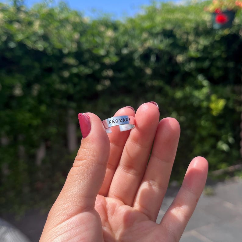 Puede incluir: Un anillo plateado con la palabra "FERRARI" grabada en la parte delantera. El anillo se sostiene contra un fondo de follaje verde y cielo azul.