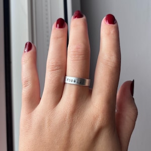 May include: A silver ring with the word "POGUES" engraved on it. The ring is worn on a person's finger.