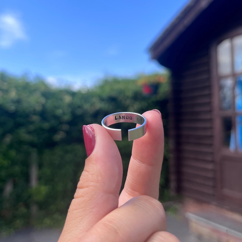May include: A silver-colored adjustable ring with the word "LANDO" engraved on the top. The ring is held up against a backdrop of a blue sky and green foliage.