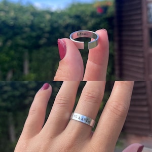 May include: Two silver rings with stamped text. One ring reads "LANDO" and the other reads "MC LAREN". The rings are displayed against a blurred green background. The rings are simple bands with a small gap.
