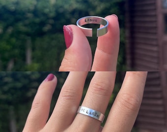 May include: Two silver rings with stamped text. One ring reads "LANDO" and the other reads "MC LAREN". The rings are displayed against a blurred green background. The rings are simple bands with a small gap.