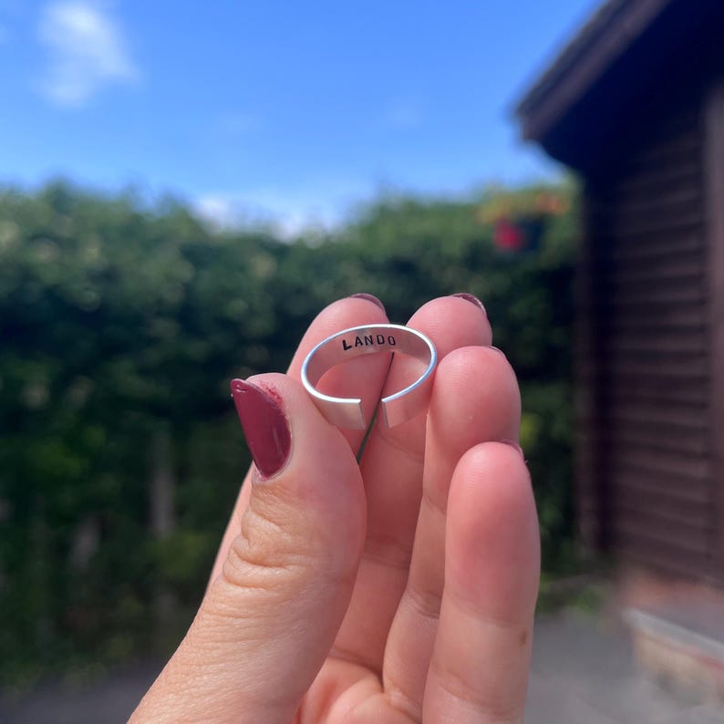 May include: A silver-colored adjustable ring with the word "LANDO" engraved on the inside. The ring is held up against a backdrop of green foliage and a blue sky.