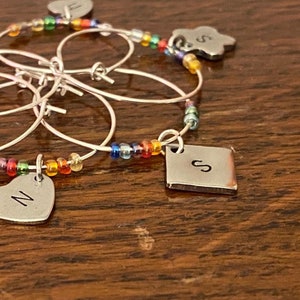 Puede incluir: Conjunto de seis charms de plata para copas de vino con cuentas de colores. Cada charm tiene una letra diferente, incluyendo 'N' y 'S'.