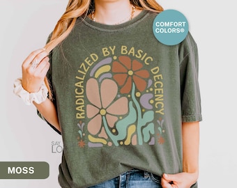 May include: Moss green t-shirt with a floral graphic and the text "RADICALIZED BY BASIC DECENCY". The design features flowers in shades of peach, orange, and purple. The shirt is a Comfort Colors brand.