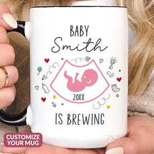 May include: White ceramic mug with a black handle. The mug has a pink ultrasound image with a baby inside and the text "Baby Smith is brewing" with a year in the middle. The mug is decorated with small drawings of baby items like a rattle, a pacifier, and a baby sock.