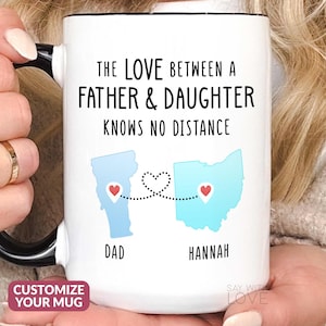 May include: White ceramic mug with a black handle and rim. The mug features the text "THE LOVE BETWEEN A FATHER & DAUGHTER KNOWS NO DISTANCE." It also shows two states connected by a dotted line with hearts, and the names "DAD" and "HANNAH."