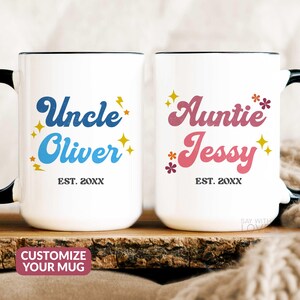 May include: Two white ceramic mugs with black handles. The mug on the left has a blue and yellow design with the text "Uncle Oliver EST. 20XX". The mug on the right has a pink and yellow design with the text "Auntie Jessy EST. 20XX".