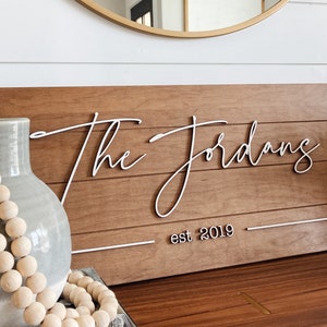 May include: A wooden sign with the text "The Jordans est 2019" in white cursive lettering. The sign is on a wooden surface with a vase and beads in the foreground.