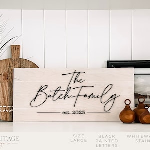 May include: A whitewash stained wooden sign with the text "The Batch Family est. 2023" in black painted letters. The sign is displayed on a white shelf with other home decor items.