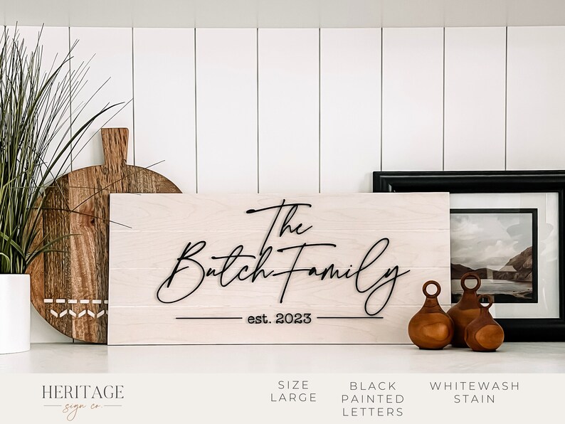 May include: A whitewash stained wooden sign with the text "The Batch Family est. 2023" in black painted letters. The sign is displayed on a white shelf with other home decor items.