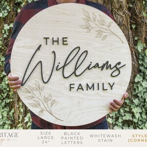 May include: A 24 inch round wooden sign with the text "THE Williams FAMILY" in black painted letters. The sign has a whitewash stain and a delicate floral design.
