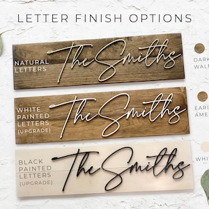 May include: Three wooden signs with the text "The Smiths" in different finishes. The top sign has natural wood with brown letters. The middle sign has a dark wood stain with white letters. The bottom sign has a light wood stain with black letters.