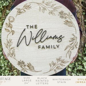 May include: A 24-inch round wooden sign with a floral wreath design. The sign says "The Williams Family" in black painted letters.