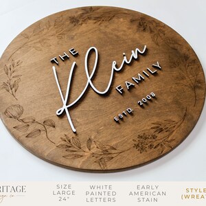 May include: A 24-inch round wooden sign with a floral wreath design. The sign features the text "The Klein Family Est. 2008" in white raised letters.
