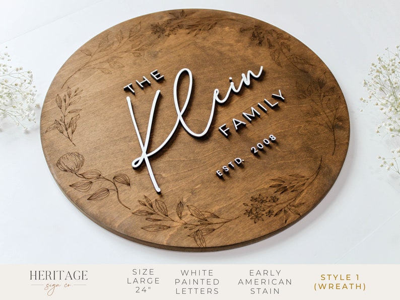 May include: A 24-inch round wooden sign with a floral wreath design. The sign features the text "The Klein Family Est. 2008" in white raised letters.
