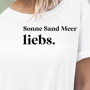 Könnte beinhalten: Weißes T-Shirt mit dem Aufdruck "Sonne Sand Meer liebs." in Schwarz. Der Text ist mittig auf dem Shirt platziert. Das Shirt ist in verschiedenen Größen und Farben erhältlich. Das Bild enthält außerdem den Text "Bügelbild" und "Verschiedene: - Größen - Farben".