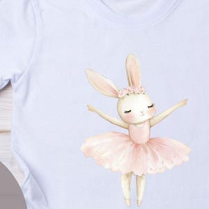 Könnte beinhalten: Weißes T-Shirt mit einer Aquarell-Illustration einer Hasen-Ballerina in einem rosa Tutu und Blumenkranz. Der Text "FERTIGES BÜGELBILD" ist auf einem grauen Kreis aufgedruckt. Das Shirt hat kurze Ärmel.