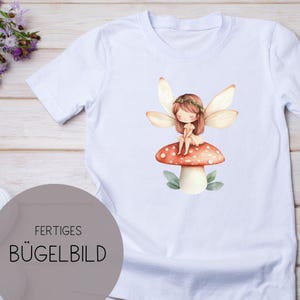 Könnte beinhalten: Weißes T-Shirt mit einer Illustration einer Fee, die auf einem rot-weiß gepunkteten Pilz sitzt. Die Fee hat braunes Haar, einen Blumenkranz und Flügel. Der Text "FERTIGES BÜGELBILD" ist unten links aufgedruckt.