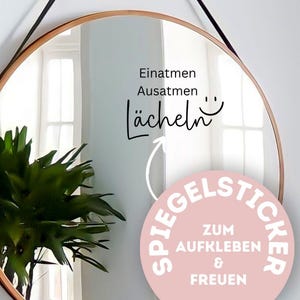 Könnte beinhalten: Ein runder Spiegel mit einem dünnen, goldfarbenen Rahmen hängt an einer weißen Wand. Der Spiegel reflektiert ein Fenster und eine grüne Pflanze. Schwarzer Text auf dem Spiegel lautet "Einatmen Ausatmen lächeln" mit einem Smiley. Ein rosa Aufkleber sagt "SPIEGELSTICKER ZUM AUFKLEBEN & FREUEN".