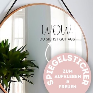Könnte beinhalten: Ein runder Spiegel mit einem dünnen, goldfarbenen Rahmen hängt an einer weißen Wand. Der Spiegel hat schwarzen Text, der lautet: "WOW. DU SIEHST GUT AUS." Ein rosa Aufkleber mit weißem Text befindet sich unten rechts.