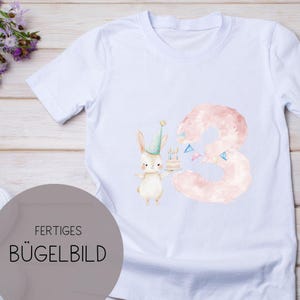 Könnte beinhalten: Weißes T-Shirt mit einer pinkfarbenen Zahl drei und einem Hasen, der einen Kuchen mit Kerzen hält. Der Text "FERtiges BÜGELBILD" befindet sich in der linken unteren Ecke des Bildes.