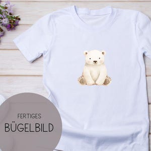 Könnte beinhalten: Weißes T-Shirt mit einer Aquarellillustration eines sitzenden Eisbären. Der Bär hat eine hellbraune Nase und Pfoten. Der Text "FERTIGES BÜGELBILD" ist auf einem grauen Kreis in der unteren linken Ecke aufgedruckt.