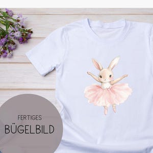Könnte beinhalten: Weißes T-Shirt mit einer Aquarell-Illustration eines Hasen-Ballerinas in einem rosa Tutu. Der Hase hat die Arme ausgestreckt und trägt Ballettschuhe. Der Text "FERTIGES BÜGELBILD" ist unten links aufgedruckt.