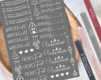 May include: A sheet of gray paper with white Christmas-themed stickers. The stickers feature various designs, including Christmas trees, ornaments, houses, stars, and text in German. The text includes "Winterzauber", "Advent", "Winterlicht", "Frohes Fest", and "Oh Tannenbaum".