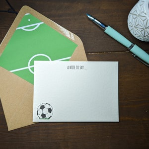 May include: A white card with a green football pitch on the front and a black and white football on the back. The card says "A note to say..." at the top.