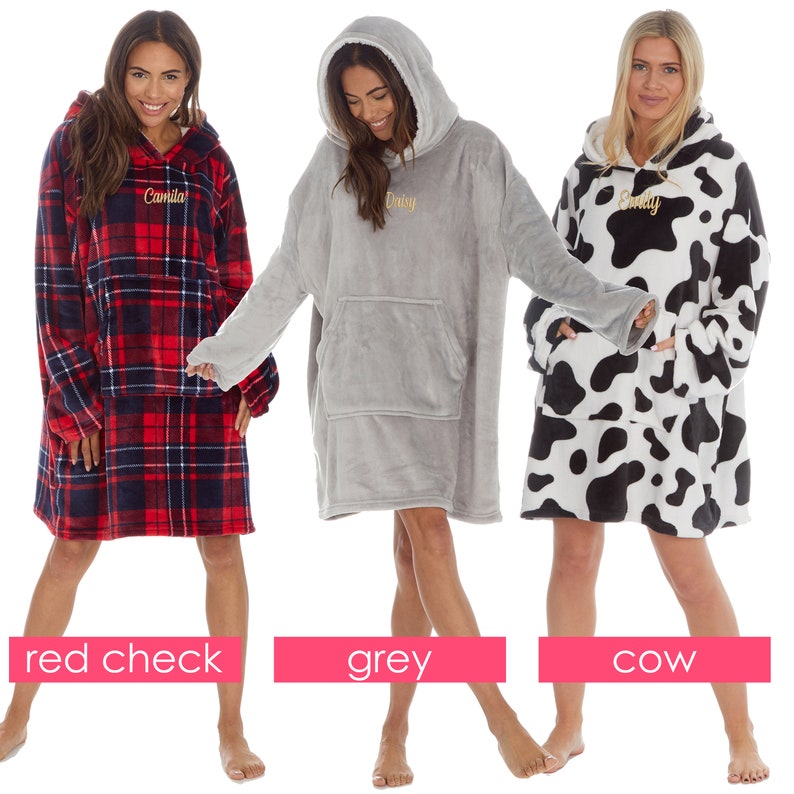 May include: Three oversized hooded blankets with different patterns. The first blanket is red and black plaid with the name "Camilla" embroidered on the front. The second blanket is grey with the name "Daisy" embroidered on the front. The third blanket is white with black cow print and the name "Emily" embroidered on the front.