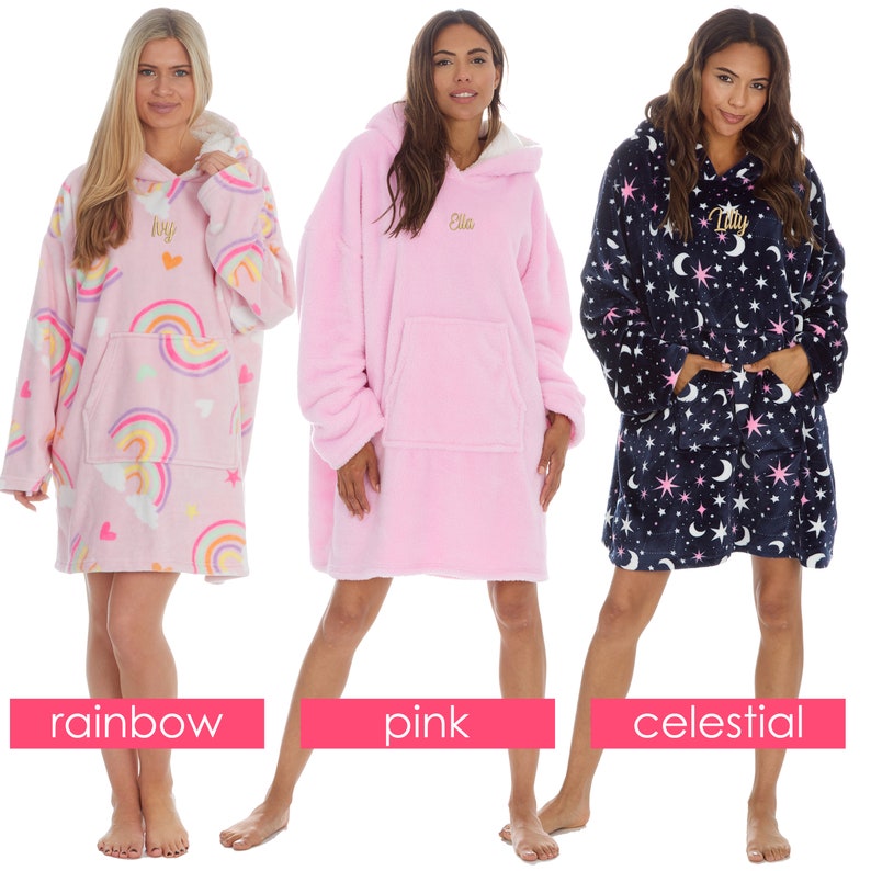 May include: Three oversized hooded blankets with different patterns. The first blanket is pink with a rainbow and heart design. The second blanket is solid pink. The third blanket is navy blue with a celestial pattern of stars and moons.
