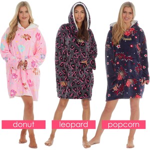 May include: Three oversized hooded blankets with different patterns. The first blanket is pink with a donut pattern. The second blanket is grey with a leopard print. The third blanket is navy blue with a popcorn pattern. Each blanket has a pocket on the front and a hood.