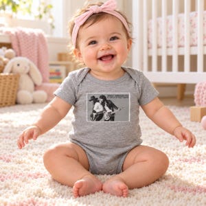 May include: A heather gray short-sleeve baby bodysuit featuring a rectangular black and white graphic print of Bruce Springsteen and the E Street Band with the text Bruce Springsteen and the E Street Band printed on the front.